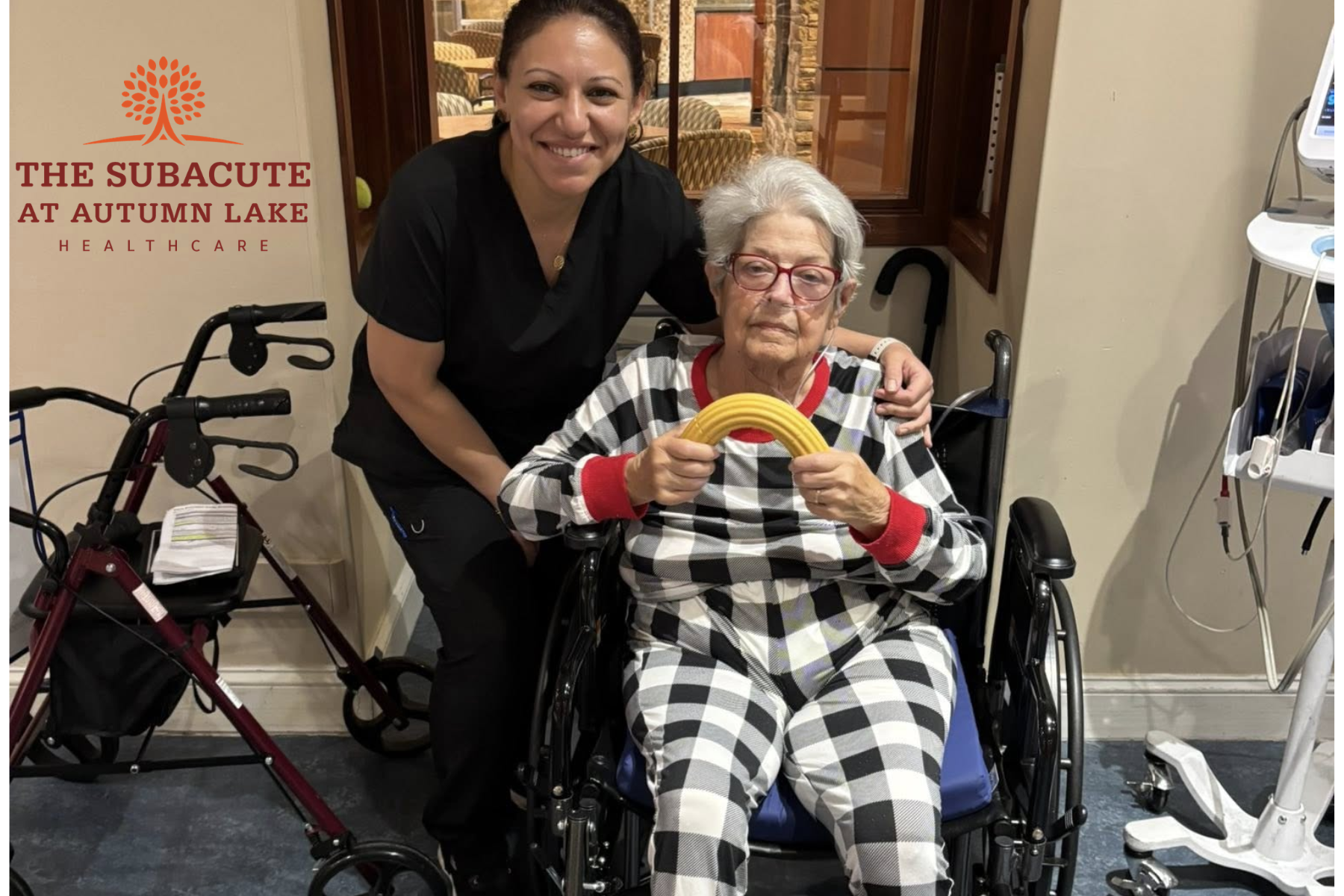 A smiling caregiver poses with an assisted living resident in a wheelchair who is holding a yellow therapeutic hand exerciser.
