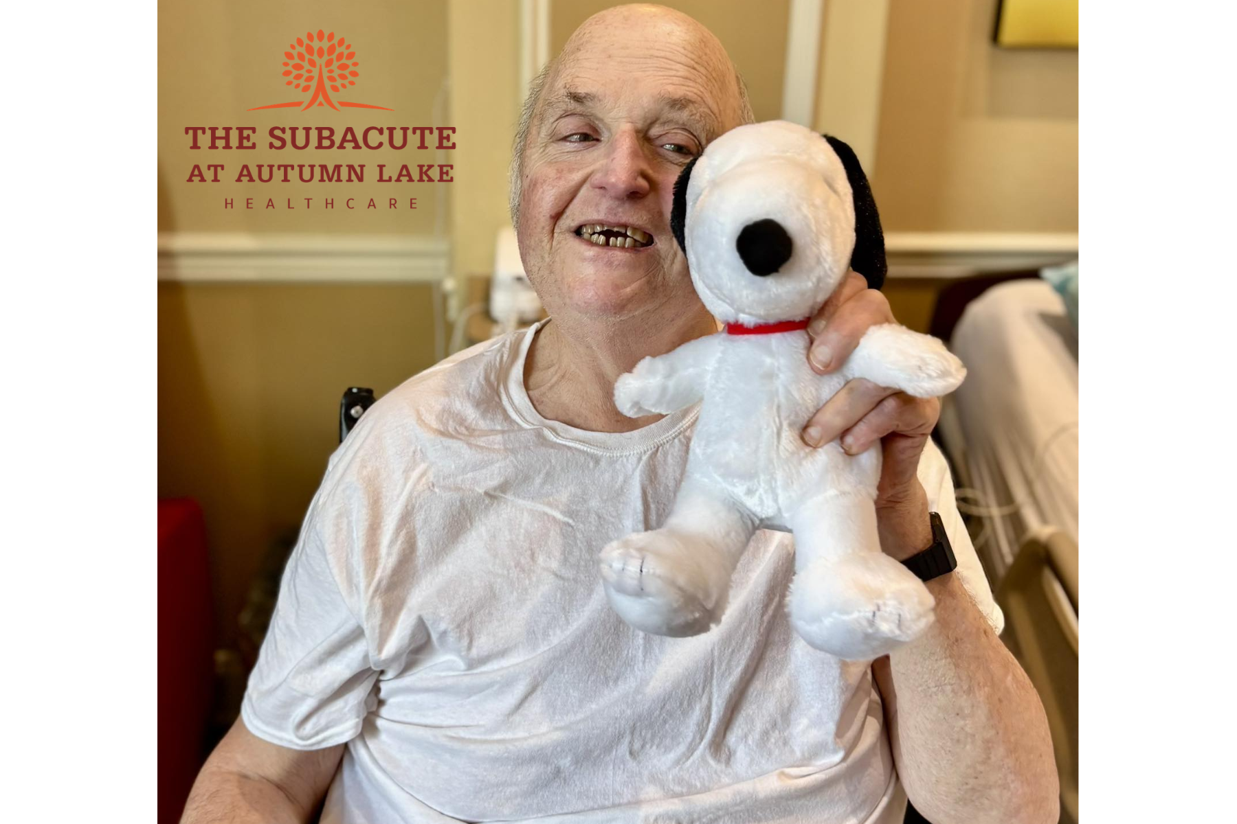 A nursing home resident smiles while holding a Snoopy plush toy while seated in his room.