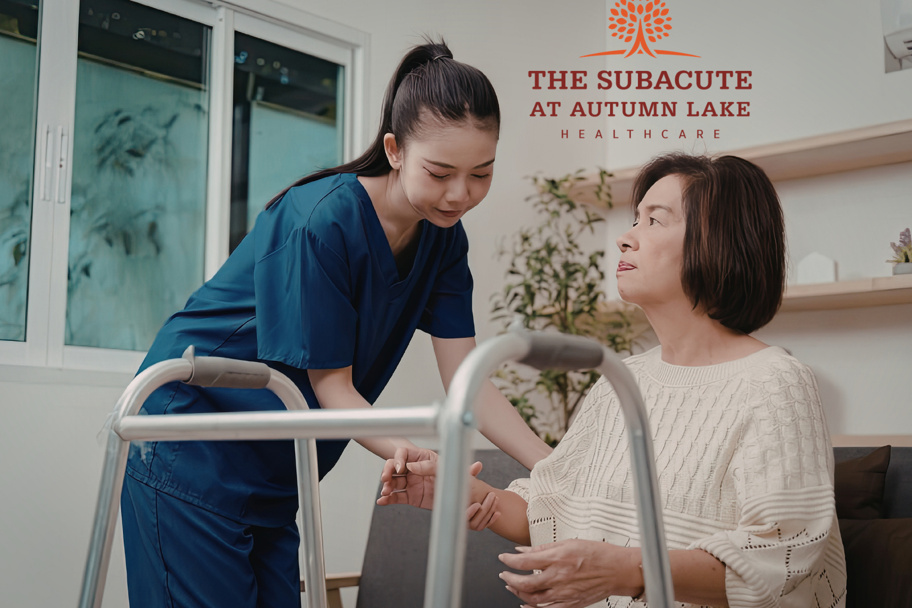 A nurse in dark blue scrubs assists an elderly woman in standing up from a couch toward her walker.